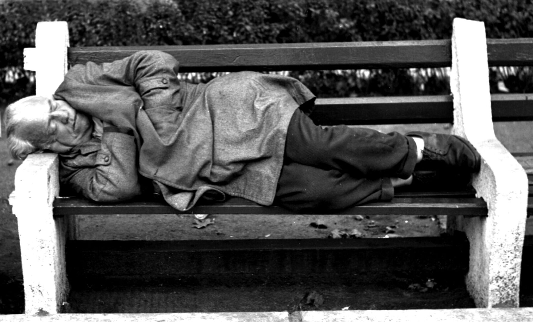 Black and white photo of a person sleeping on a bench outdoors.