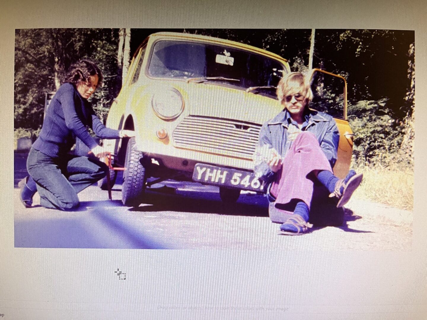 Two men sitting beside a tilted car on a street.