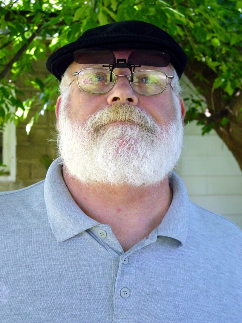 Older man with white beard and glasses outdoors under greenery.