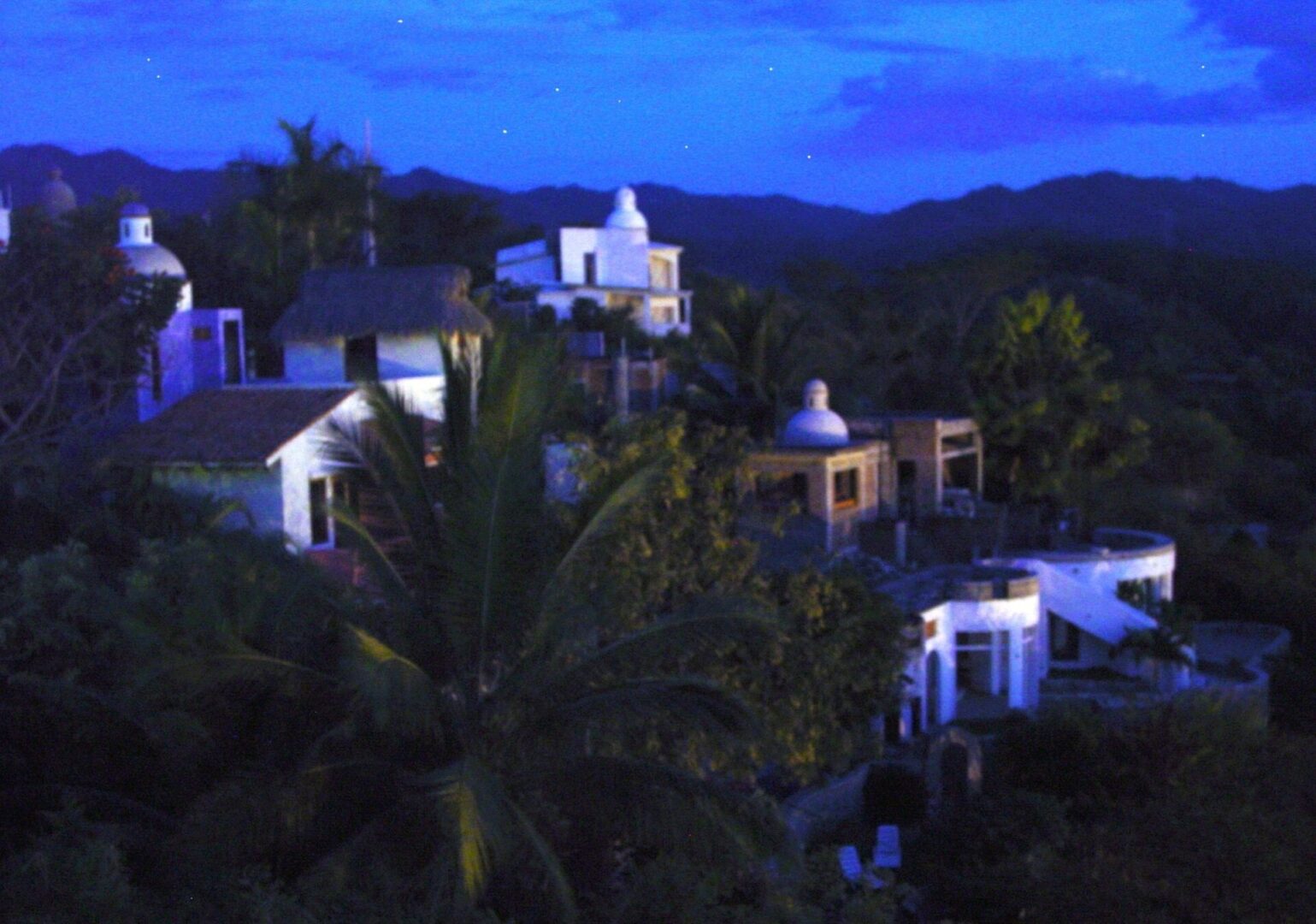 Village at dusk with illuminated white buildings and lush greenery.