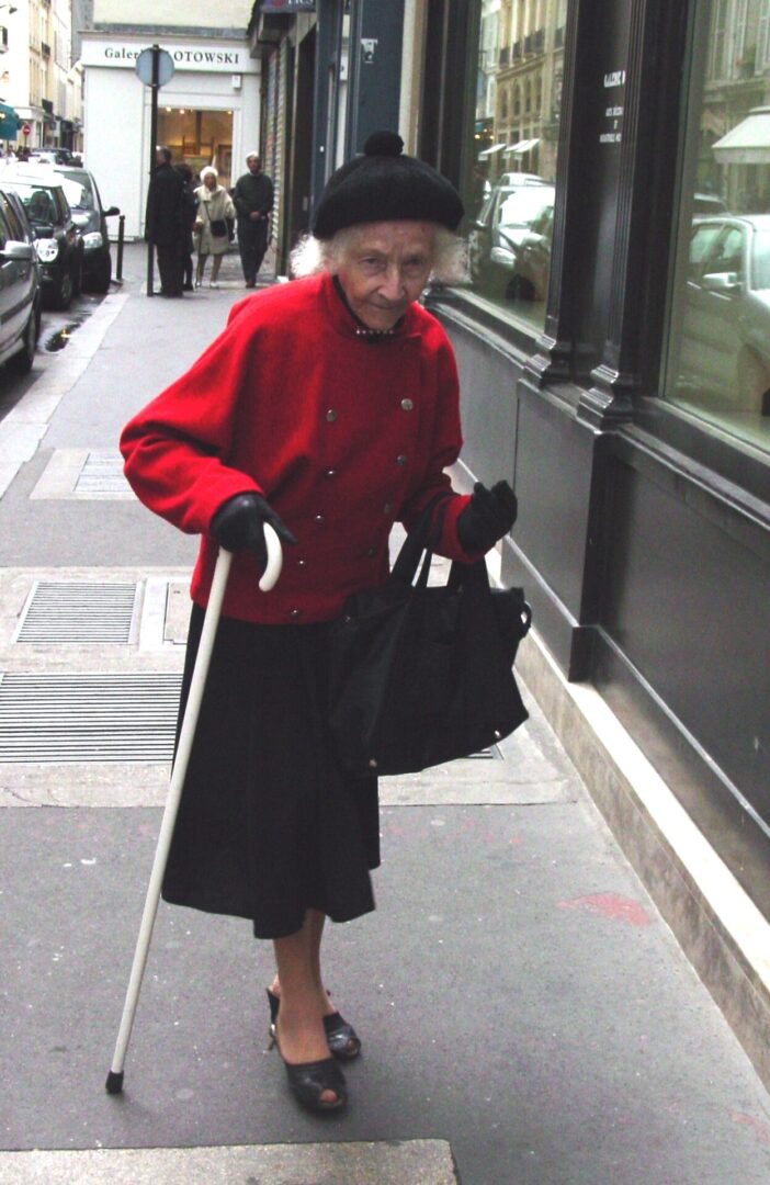 Elderly woman in red coat walking with a cane on a city sidewalk.