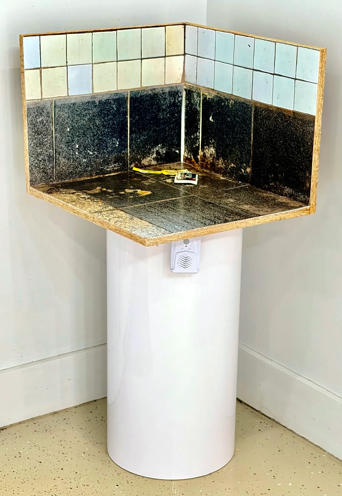 A corner pedestal sink with black tiled backsplash and white cylindrical base.