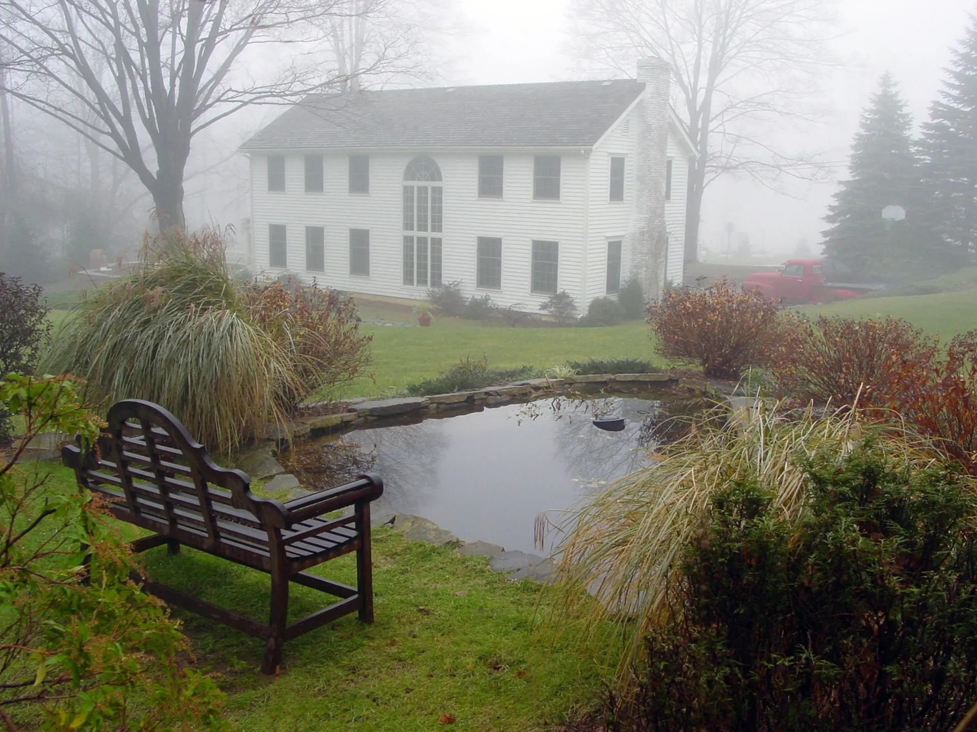 Foggy morning by a peaceful pond with a bench and white house.