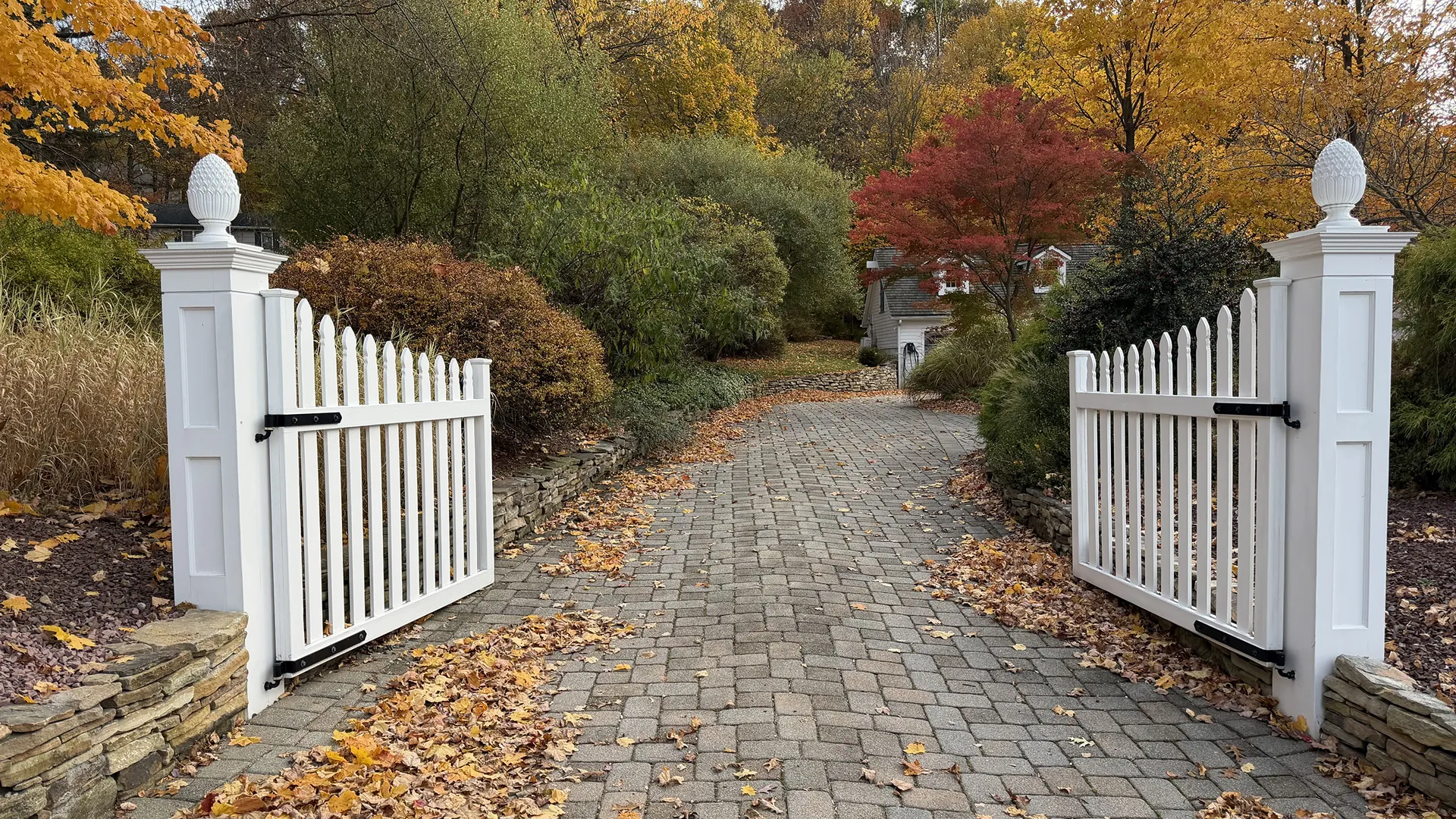 A cobblestone path through an open white gate surrounded by autumn foliage.