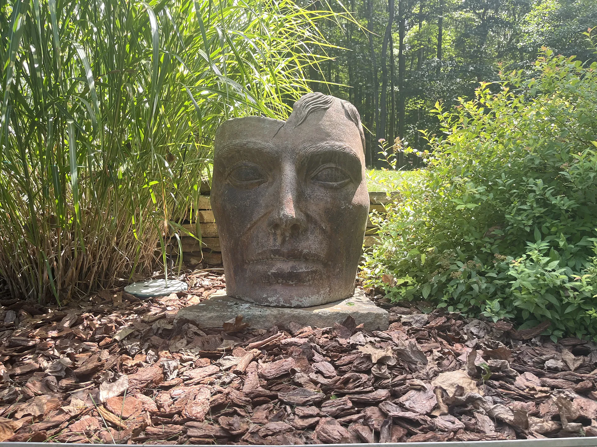 Large stone sculpture of a human face partially buried in leaves outdoors.