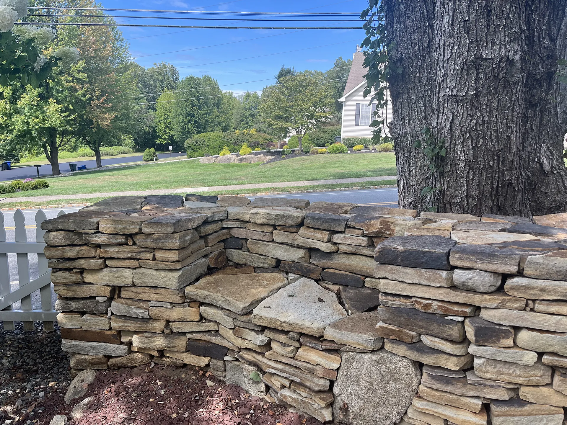 A stone wall with an uneven gap near a large tree in a suburban area.
