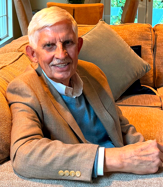 Elderly man in a brown blazer smiling while seated on a couch.